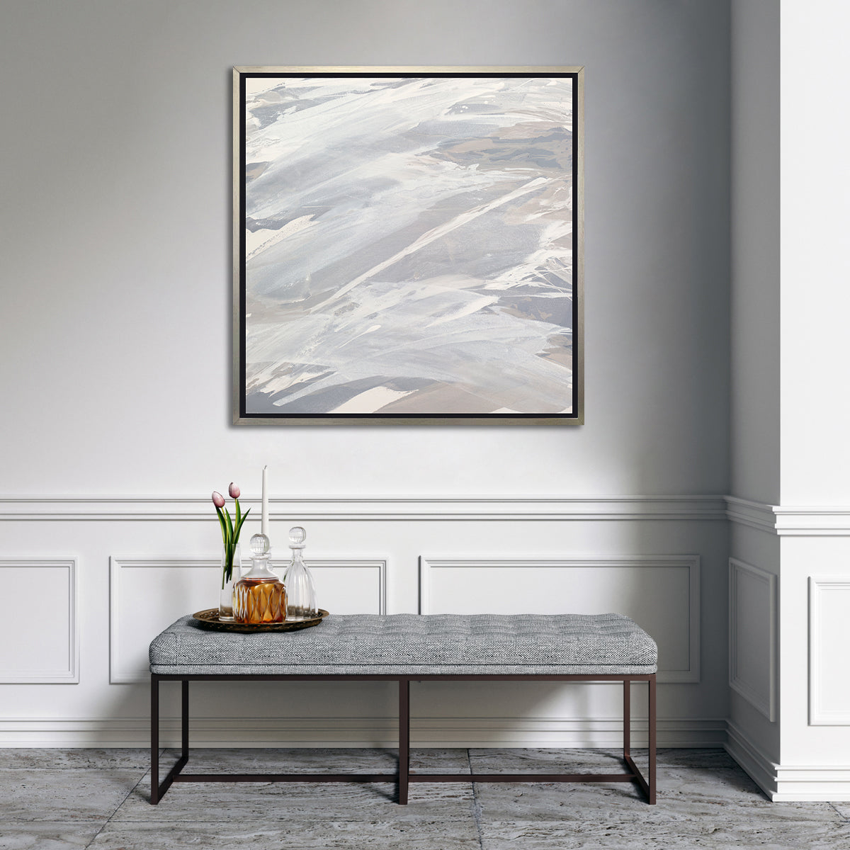 A grey, beige and white abstract print in a silver floater frame hangs on a white wall above a grey tufted bench with a tray of two glass decanters, a white tapered candlestick and a glass vase with pink tulips.