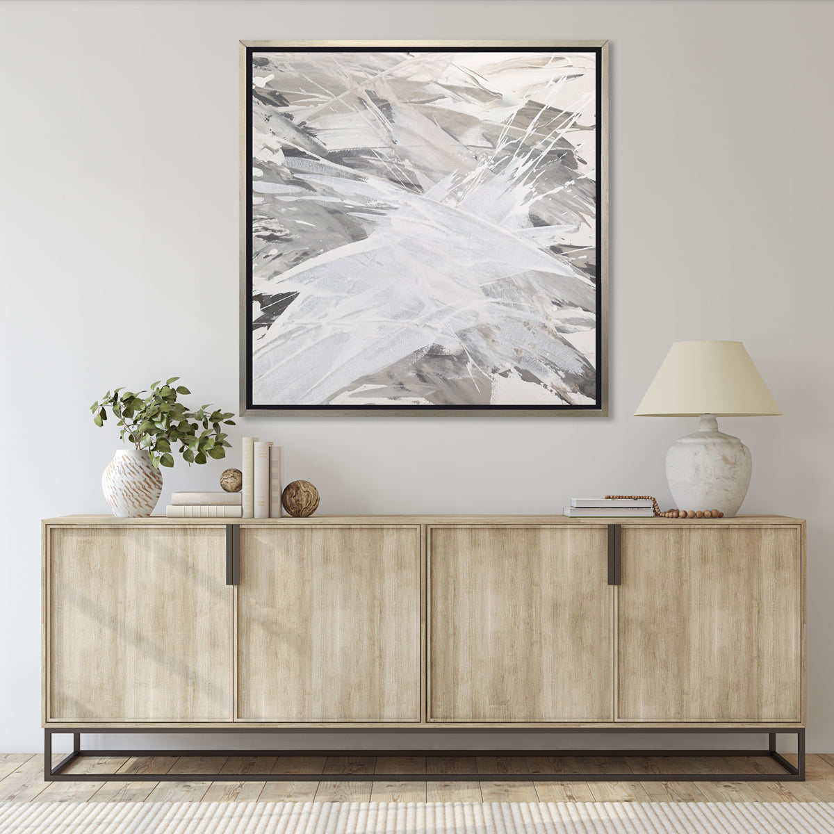 A grey, beige and white abstract print in a silver floater frame hangs on a white wall above a natural wood credenza with a stack of books, a white ceramic table lamp and a white and tan vase filled with eucalyptus leaves.
