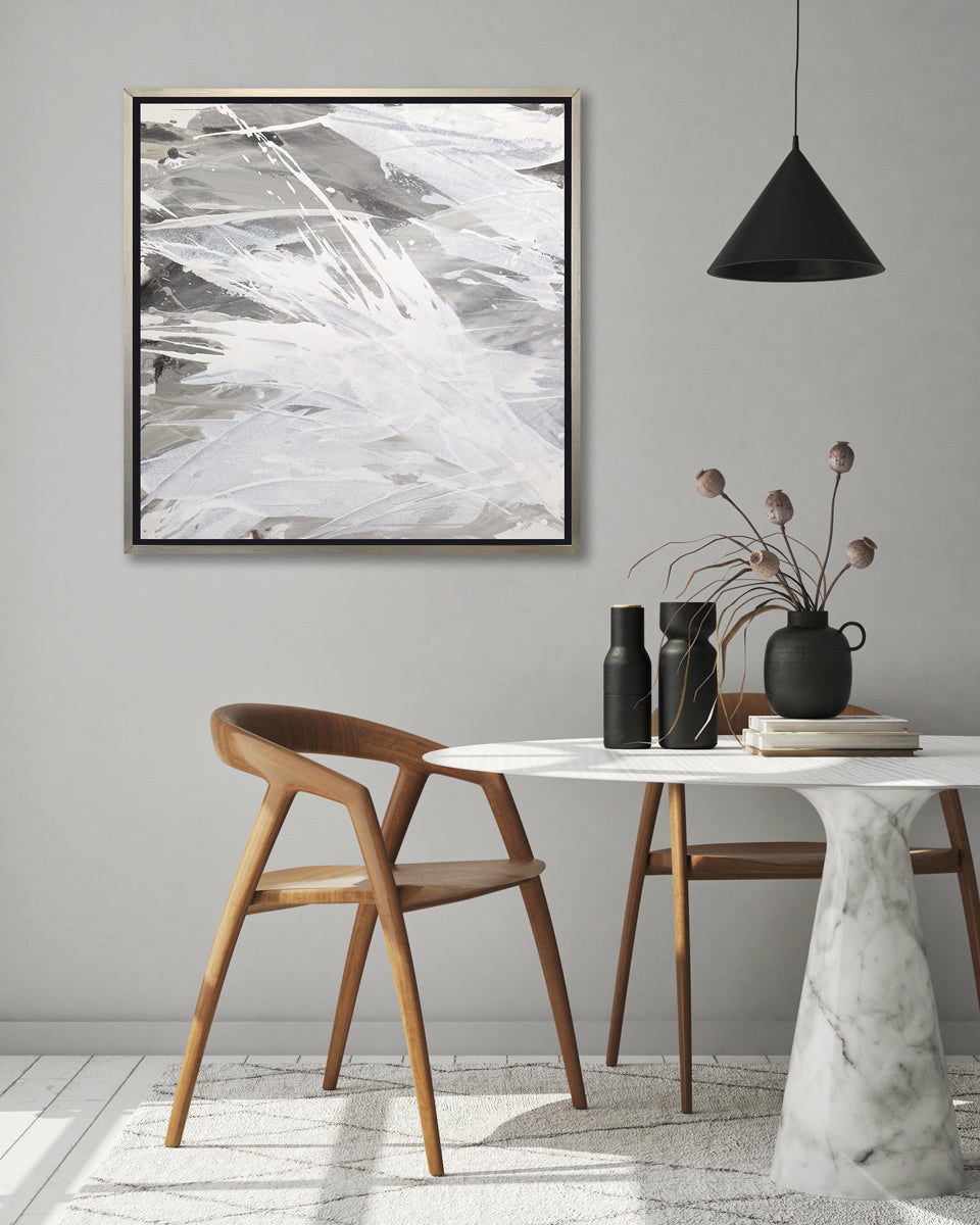 A grey, beige and white abstract print in a silver floater frame hangs on a grey wall above a white marble pedestal table with black vases and two solid wood modern chairs.