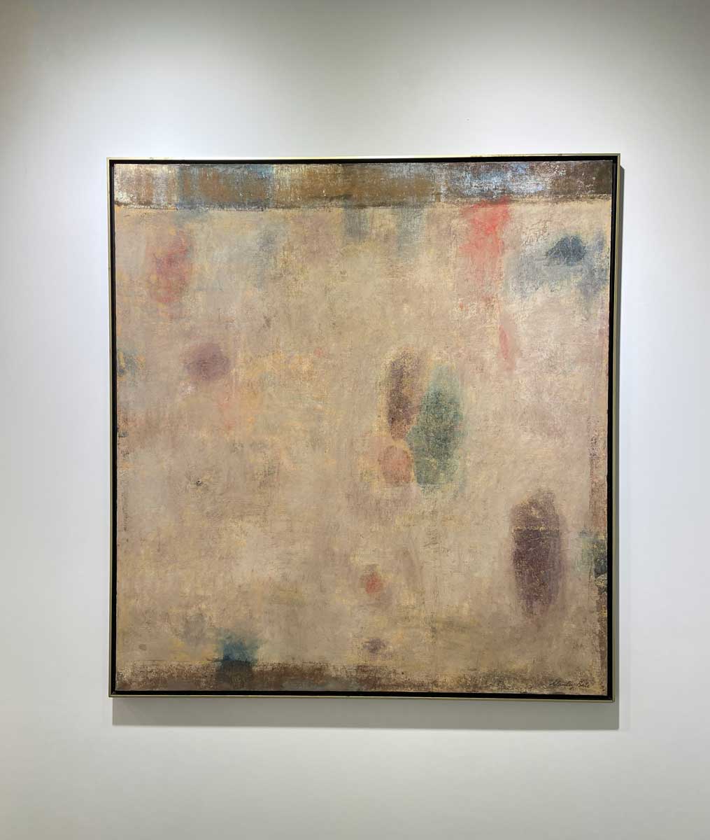 A beige painting with green, blue and purple accents in a warm gold floater frame hangs on a white wall.