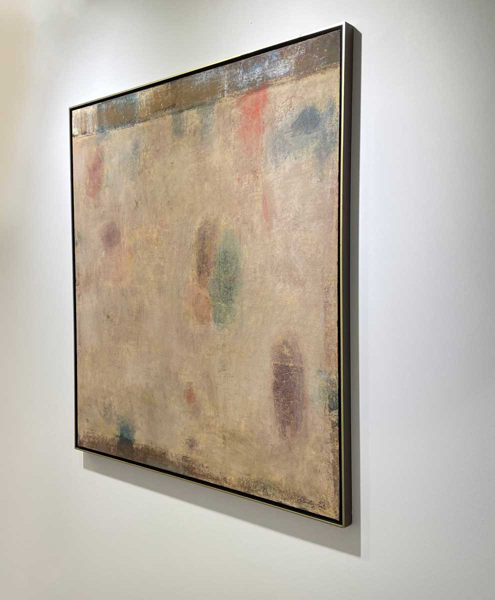 A right side-angle view of a beige painting with green, blue and purple accents in a warm gold floater frame hangs on a white wall.