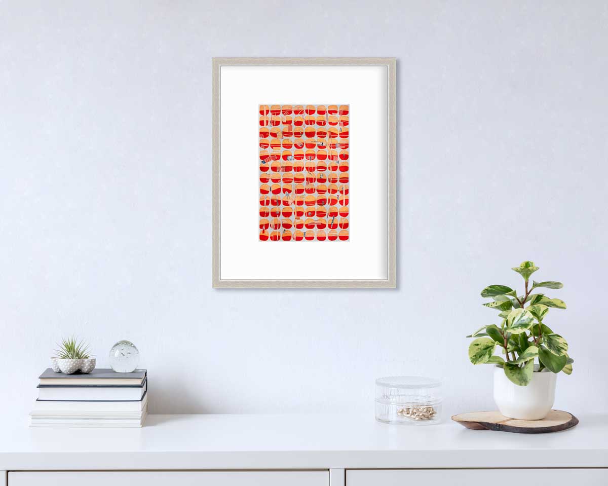 A red, orange and light blue abstract print in a silver frame with a mat hangs on a white wall above a stack of books, a round clear container with gold accessories and a potted plant.