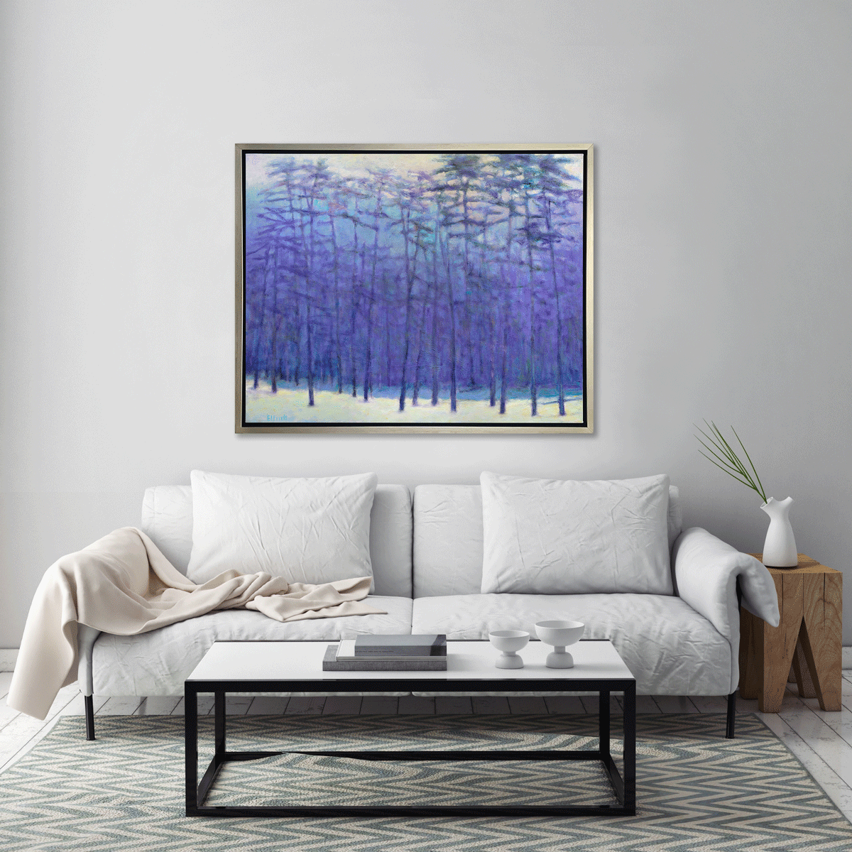 A blue, purple, light pink and soft yellow impressionistic landscape print in a silver floater frame hangs on a white wall above a white sofa with pillows, a white and black coffee table with a stack of books and a natural wood side table with a white vase.
