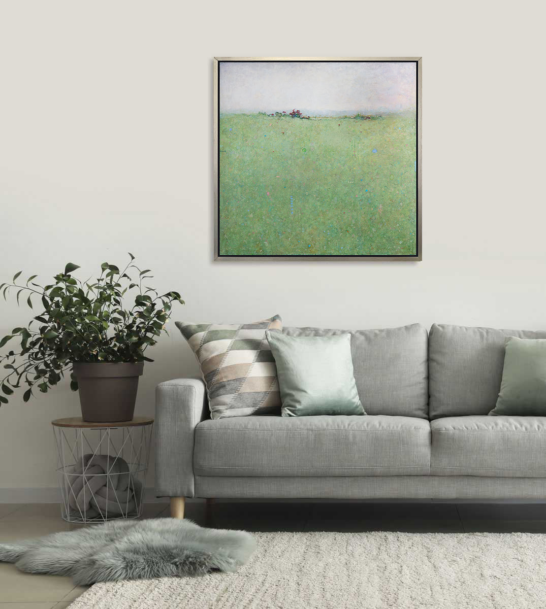 A green abstract landscape print in a silver floater frame hangs on a white wall above a grey sofa with pillows, a side table with a potted plant and a two rugs overlapping on the floor.