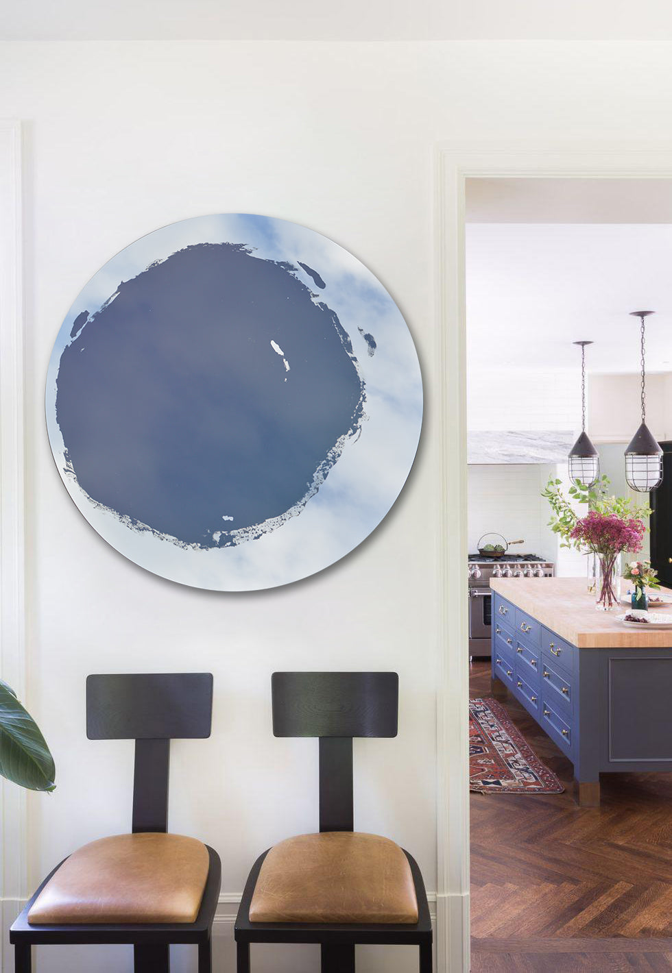 Circular mirror, mounted on wall, with blue mixed media painting in the center. Displayed on wall to the left of kitchen entryway. Chairs placed below the mirror.