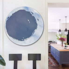 Circular mirror, mounted on wall, with blue mixed media painting in the center. Displayed on wall to the left of kitchen entryway. Chairs placed below the mirror. 