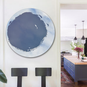 Circular mirror, mounted on wall, with blue mixed media painting in the center. Displayed on wall to the left of kitchen entryway. Chairs placed below the mirror.