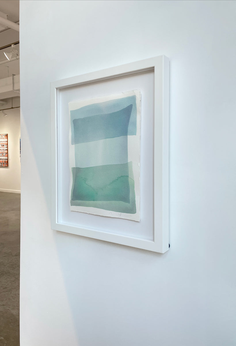 A right side-angle view of tonal blues and green shapes overlap in this painting by Nealy Hauschildt in a white frame floating above a white matte hangs on a white wall.
