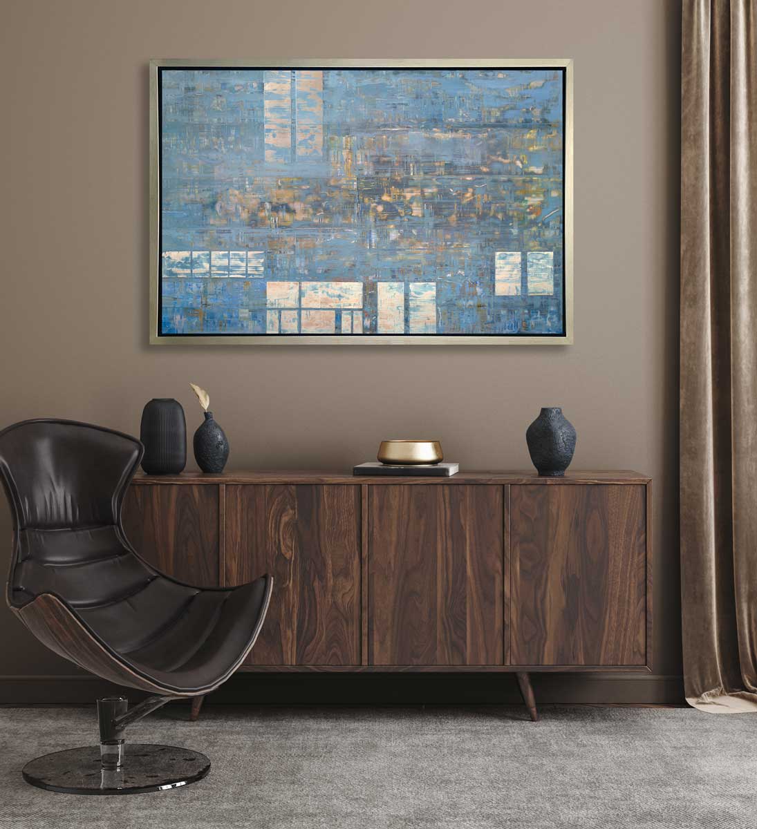 A blue and grey geometric abstract print in a silver floater frame hangs on a tan wall above a wooden credenza with black vases and a modern dark brown chair.