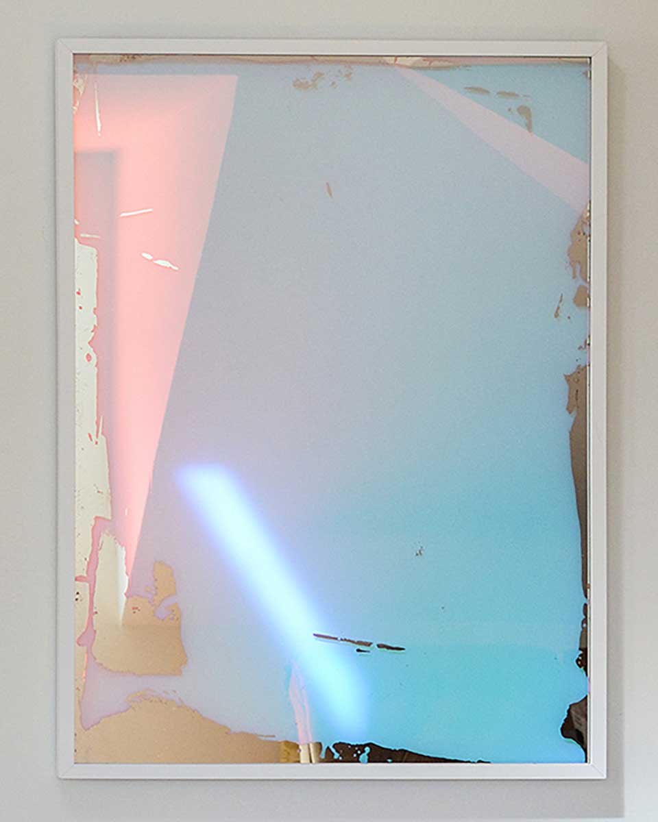 A multicolored fine art mirror framed in a white frame hangs on a white wall.
