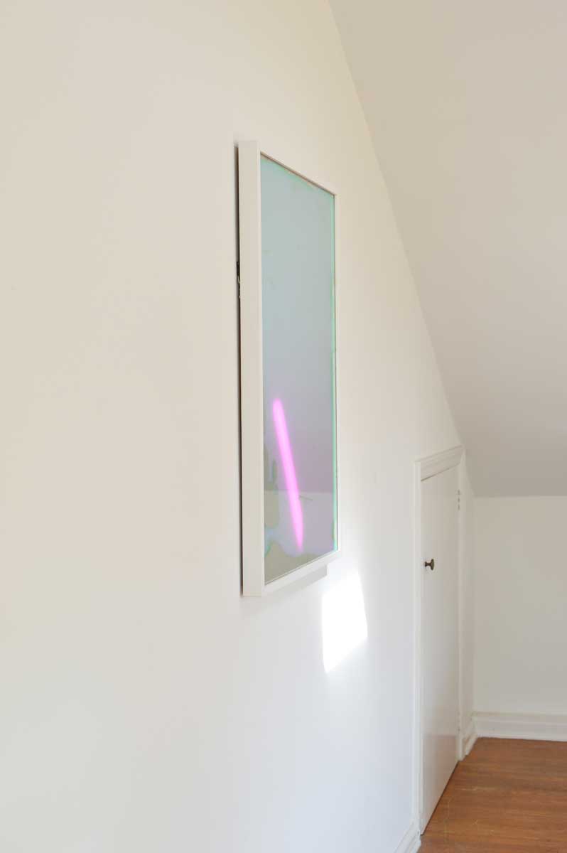 A multicolored fine art mirror hangs on a white wall near corner with a steep ceiling.