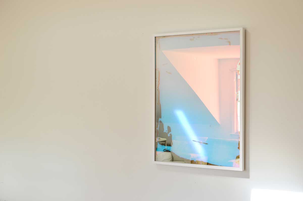 A right-hand side-angle view of a pink and blue fine art mirror hangs on a white wall, reflecting the furniture in the room.