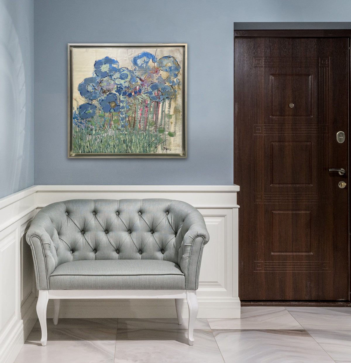 An abstract, encaustic floral painting of blue hydrangeas by Linda Bigness hangs on a blue grey wall in a warm silver floater frame above a grey armchair to the right of a walnut wood front door.