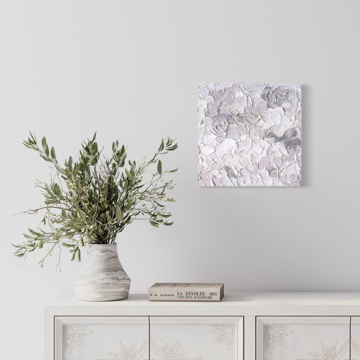 A cool neutral painting hang over a dresser. On the dresser is a marble vase with some green foliage.
