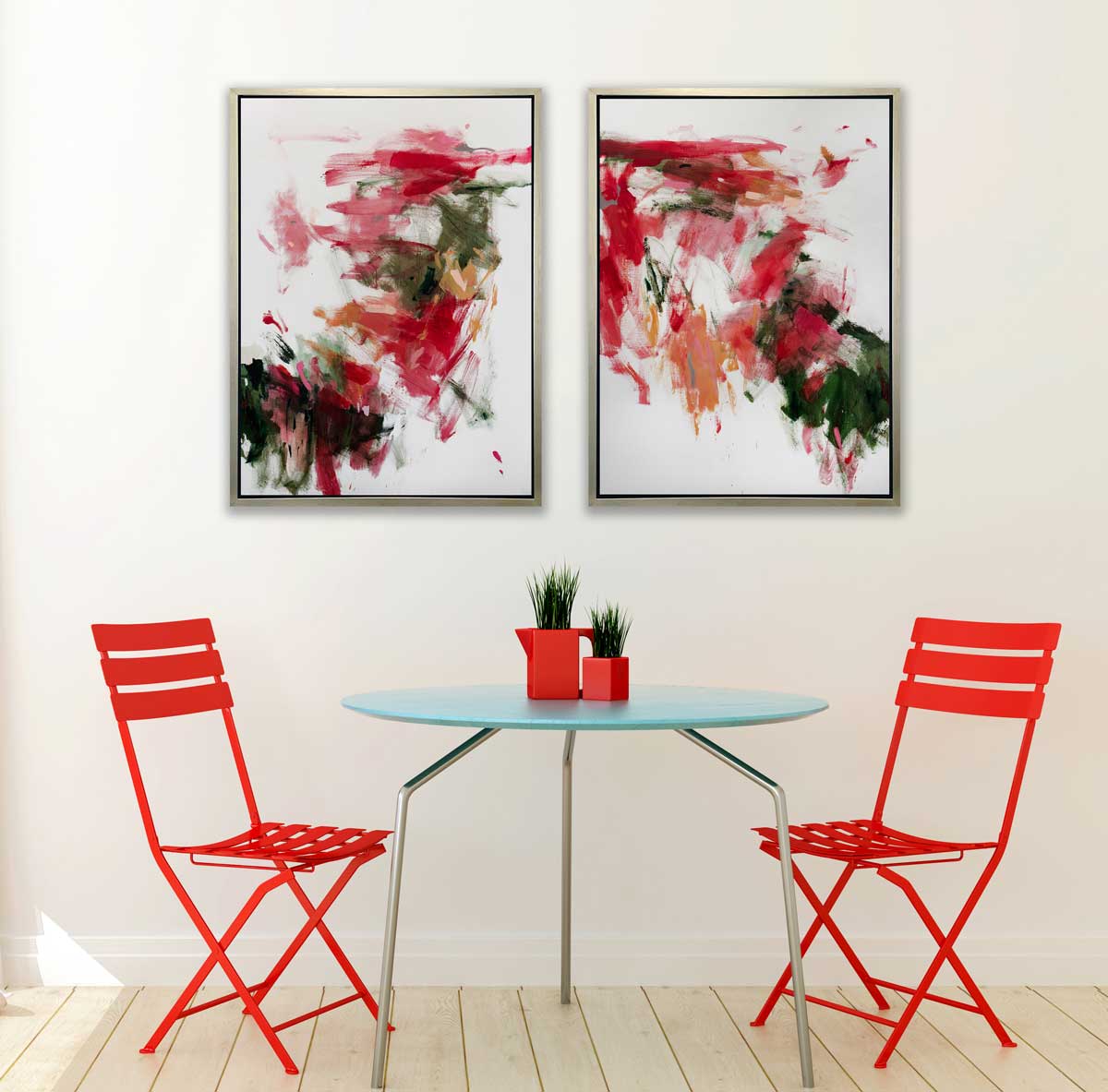 Two red, green and white abstract prints in silver floater frames hang side by side above a round teal table with red vases and greenery and two red folding chairs.