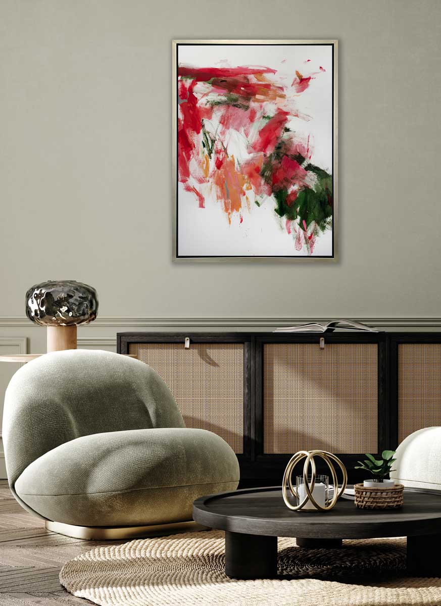 A red, green and white abstract print in a silver floater frame hangs on a light green wall above a black and rattan cabinet, a low black round coffee table and a modern pale green chair.