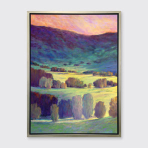 A pink, blue and green abstract landscape print in a silver floater frame hangs on a white wall.