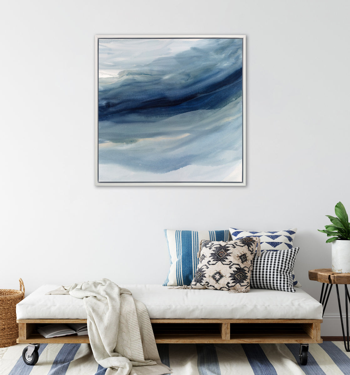 A blue, navy and white abstract print in a white floater frame hangs on a white wall above a low wheeled bench with a white cushion and blue printed pillows, a small wood side table and a woven basket.