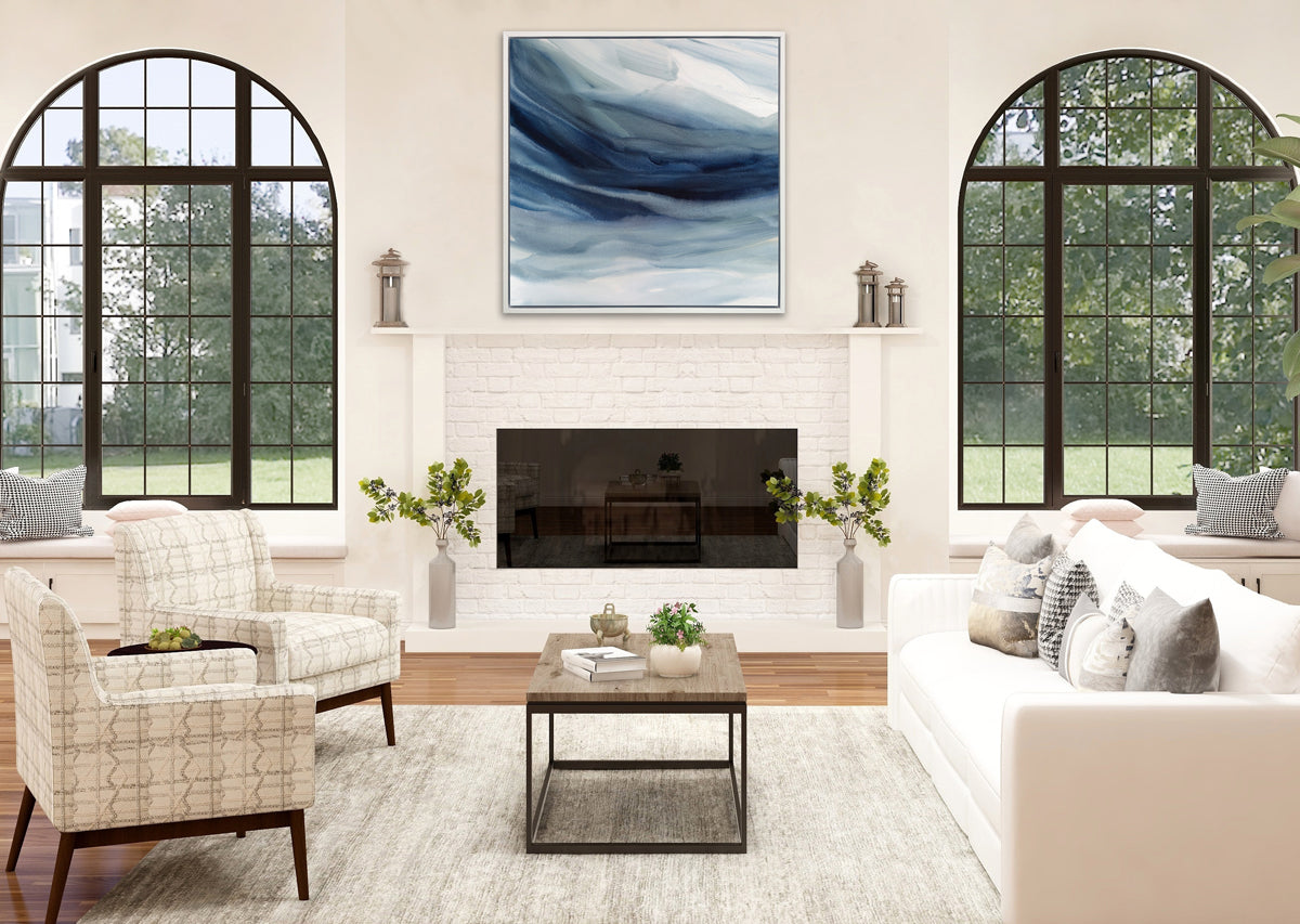 A blue, navy and white abstract print in a white floater frame hangs on a white wall in between two large arched windows and above a white brick fireplace, a large white sofa with pillows, a rectangular wood coffee table with accessories and two matching white printed accent chairs.