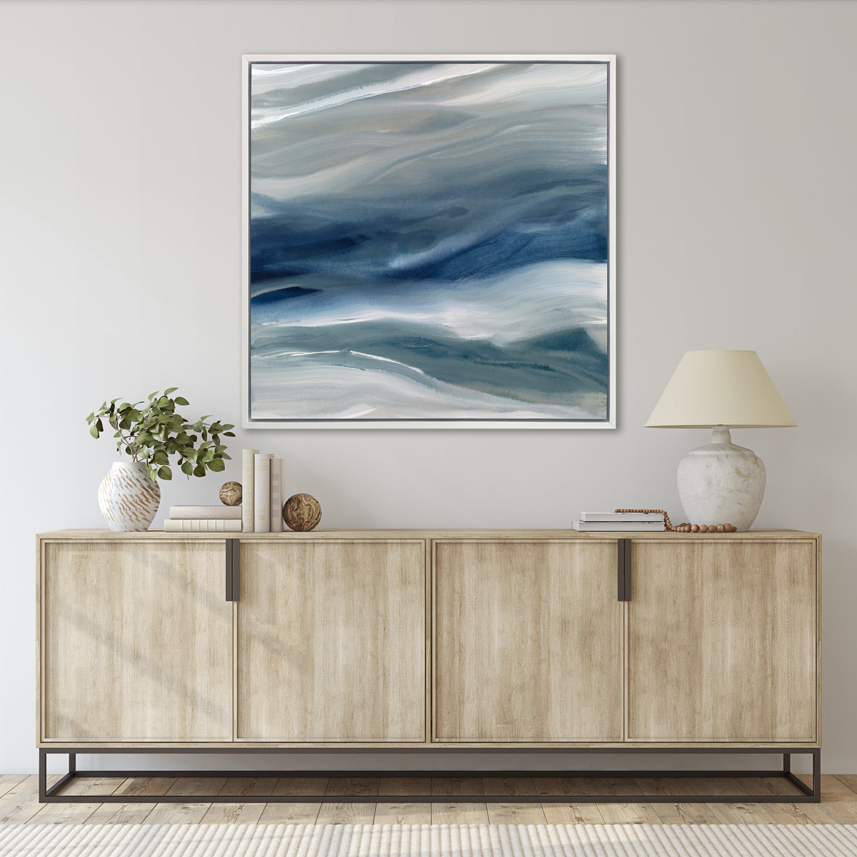 A blue, teal, beige and white abstract print in a white floater frame hangs on a white wall above a natural wood credenza with stacks of books, a marble table lamp and a white and beige vase with eucalyptus stems.
