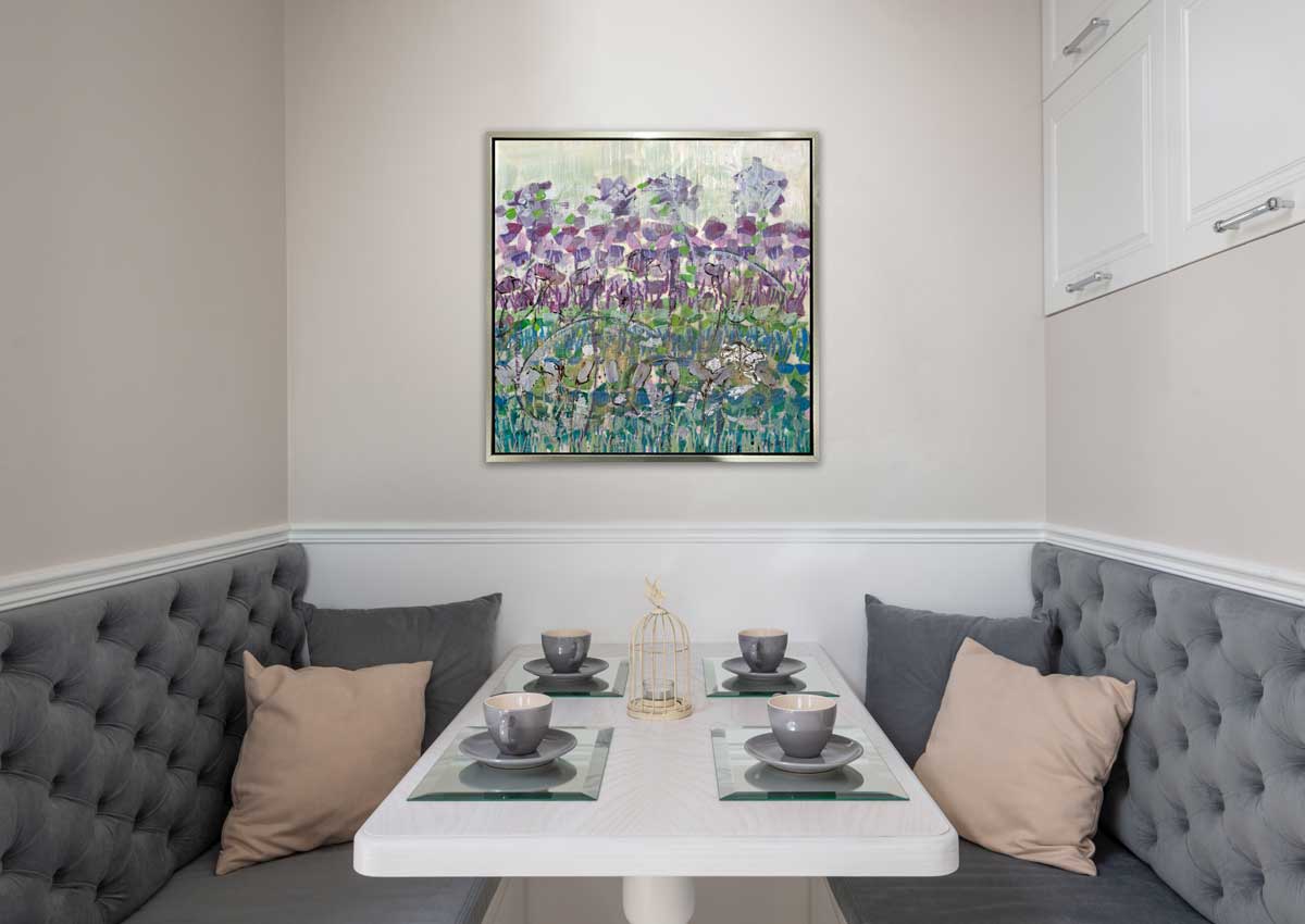 A purple floral print in a silver floater frame hangs on a white wall above a small booth with grey cushions and pillows, a white table with four place settings of grey teacups and saucers and a small off-white birdcage tealight holder in the center of the table.