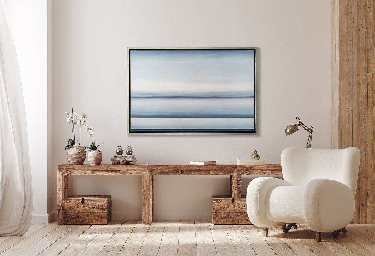 A tonal blue abstract landscape print in a silver floater frame hangs on a white wall above a long natural wood desk with two potted white orchids, a stack of books and a chrome adjustable desk lamp. A white modern armchair sits to the right.