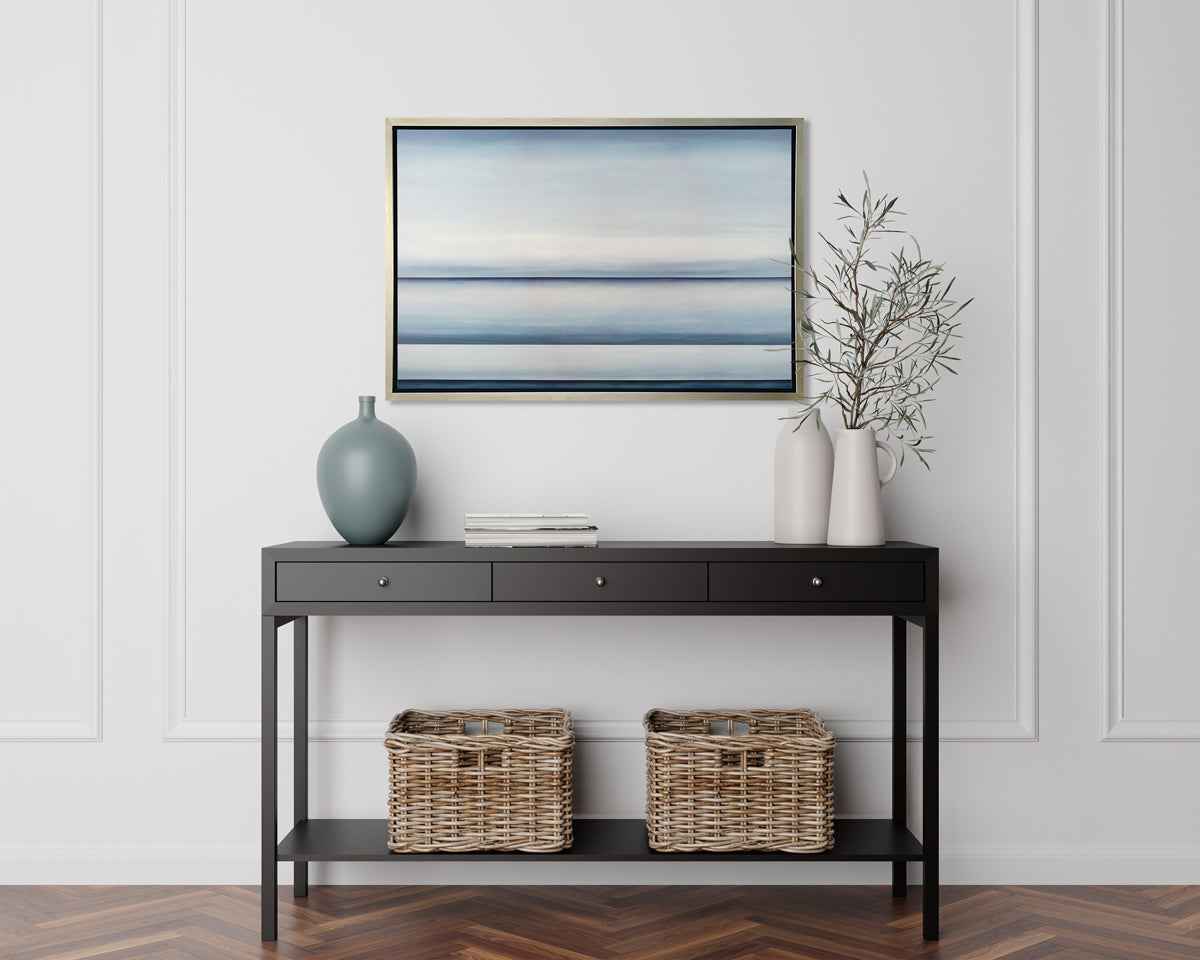 A tonal blue abstract landscape print in a silver floater frame hangs on a white wall above a black console table with a muted teal vase, a stack of books and two white pitchers, one full of greenery stems.