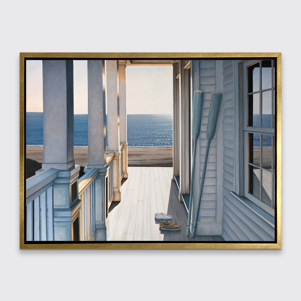 A print of a sunlit porch overlooking the water with two oars and two pairs of shoes on the deck in a gold floater frame hangs on a white wall.