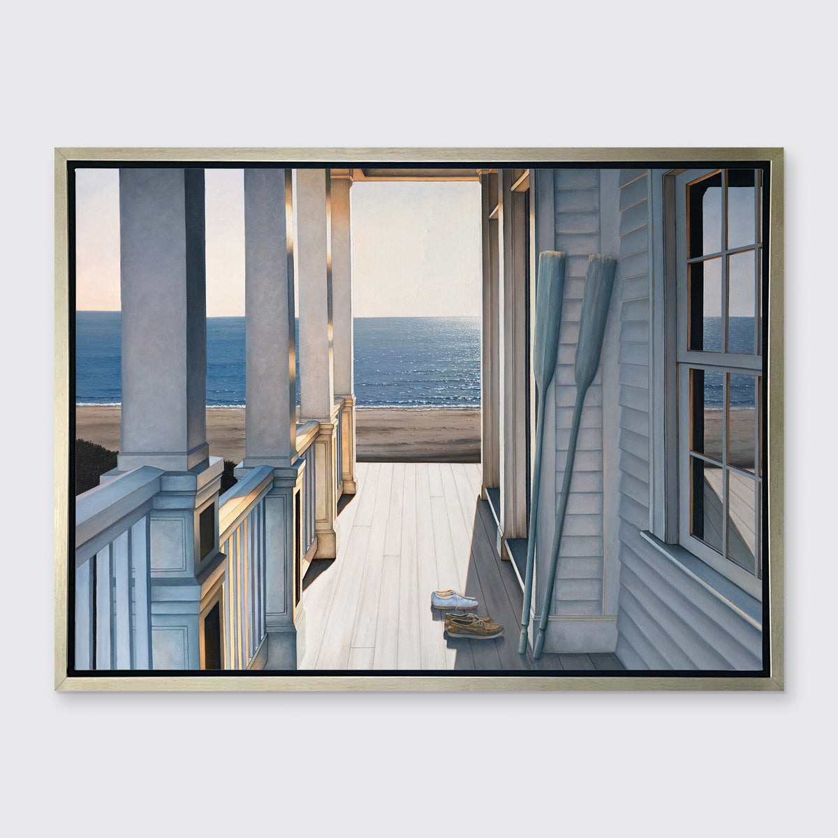 A print of a sunlit porch overlooking the water with two oars and two pairs of shoes on the deck in a silver floater frame hangs on a white wall.
