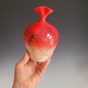 A hand holds a red ceramic vessel in front of a grey gradient backdrop. 