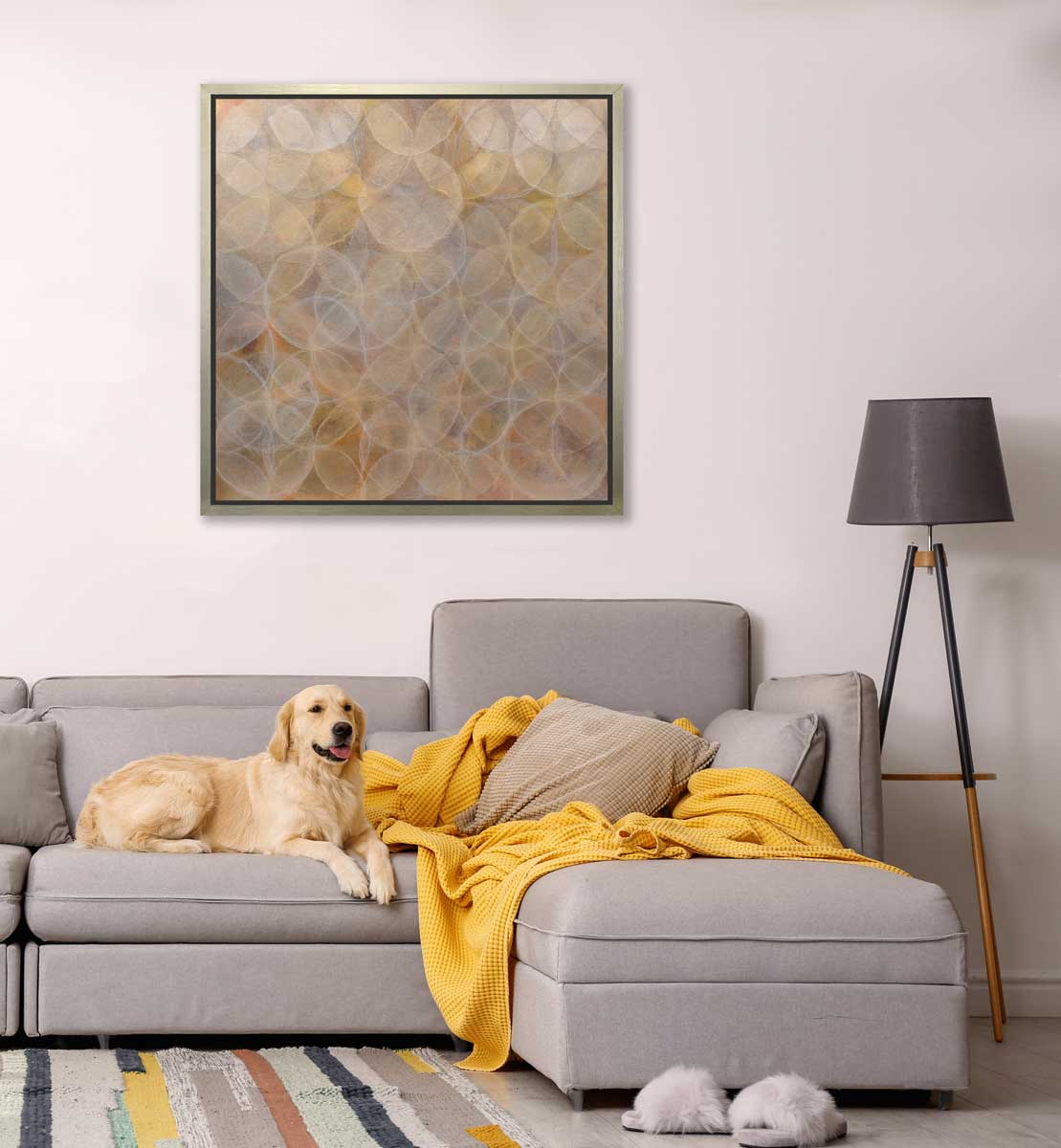 A dark yellow and white abstract overlapping circle print in a silver floater frame hangs on a white wall above a grey sofa with a large yellow throw blanket, a golden retriever laying down and a tripod floor lamp.