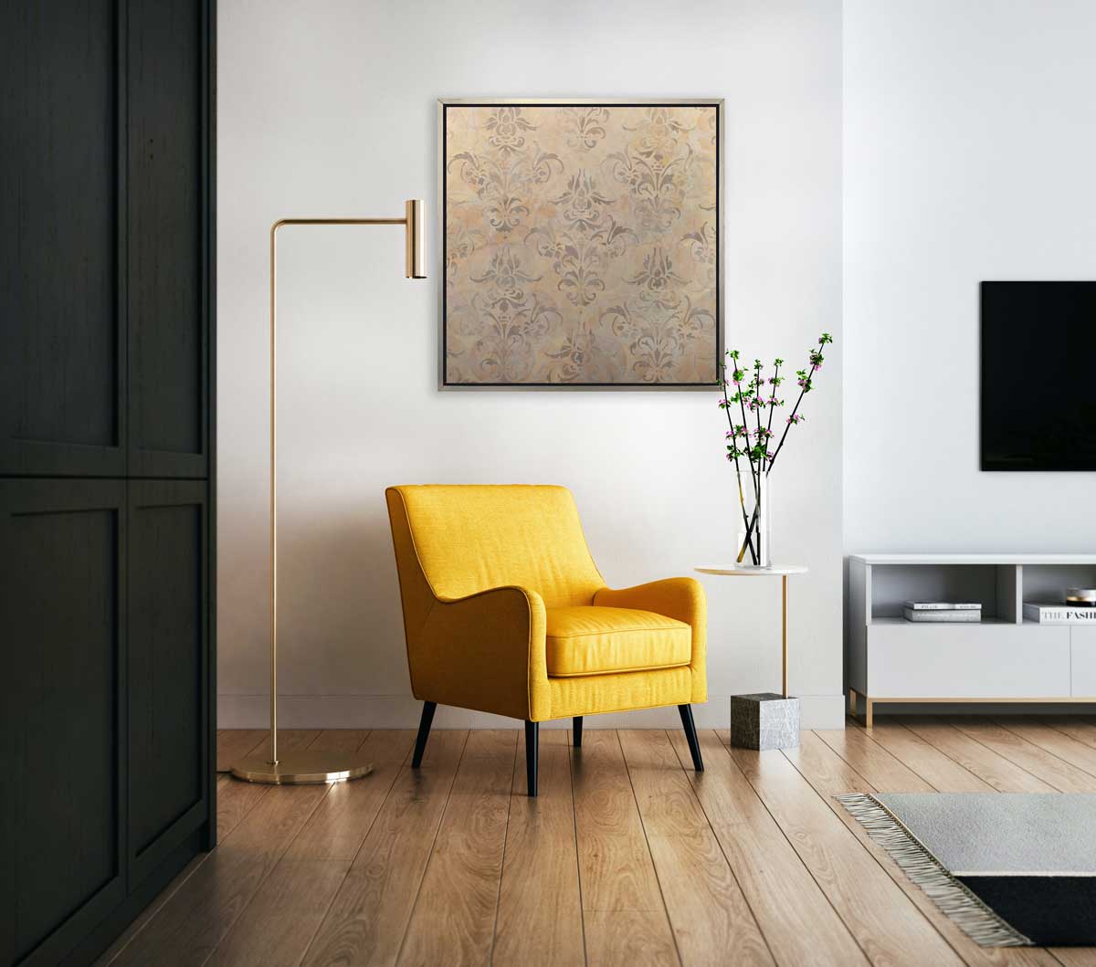 A light gold and silver abstract print in a silver floater frame hangs on a white wall above a yellow chair, a gold floor lamp and a side table with a clear vase full of purple flowers.
