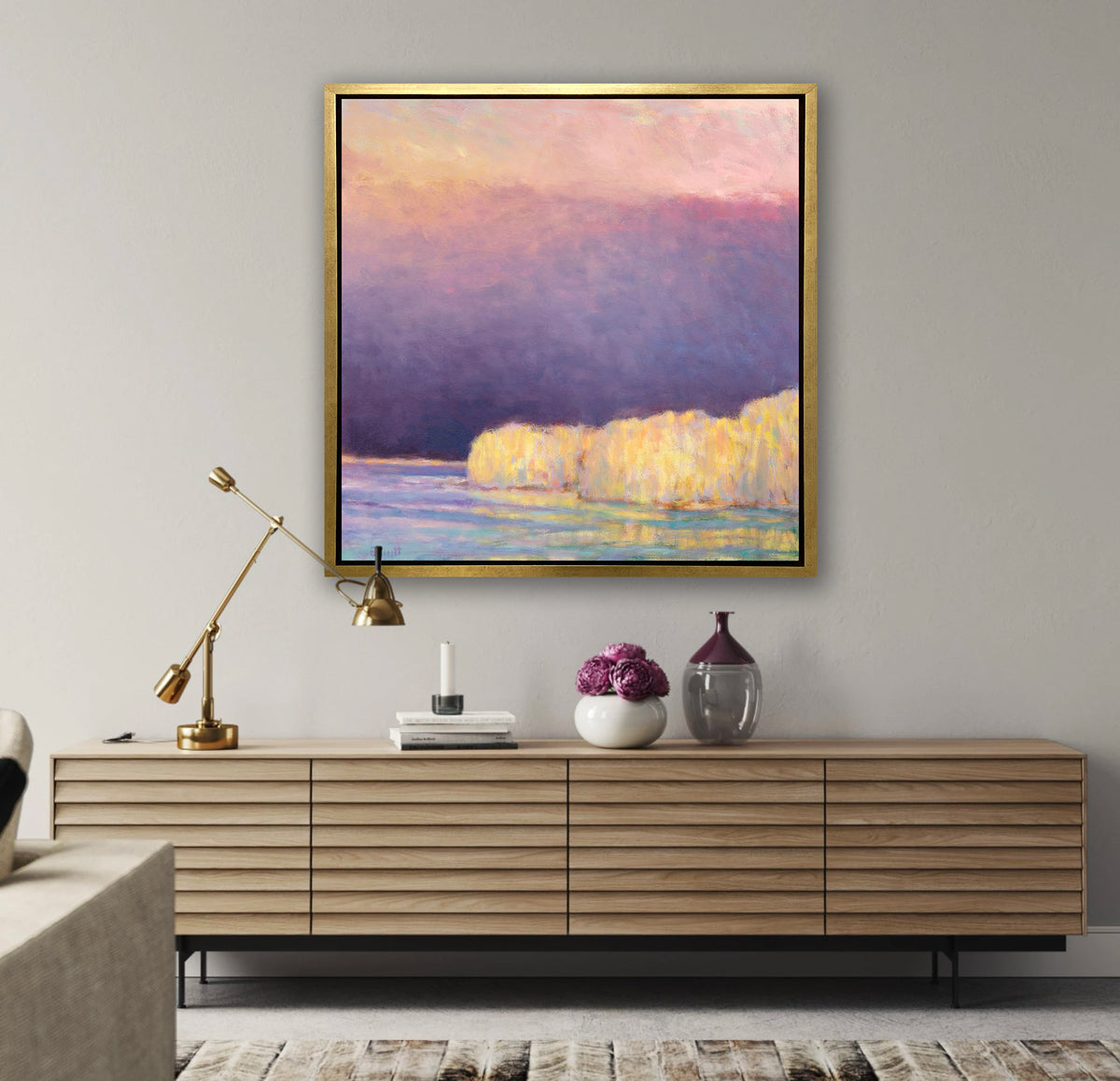 A violet, pink, yellow and light blue abstract landscape print in a gold floater frame hangs on a white wall above a long wood console table with an adjustable gold lamp, a stack of books and two vases with violet flowers.
