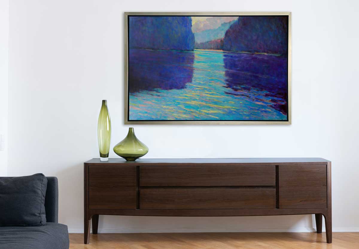 A dark blue and teal abstract landscape print in a silver floater frame hangs on a white wall above a dark brown console table with two green vases and a partial section of a denim sofa with a pillow.