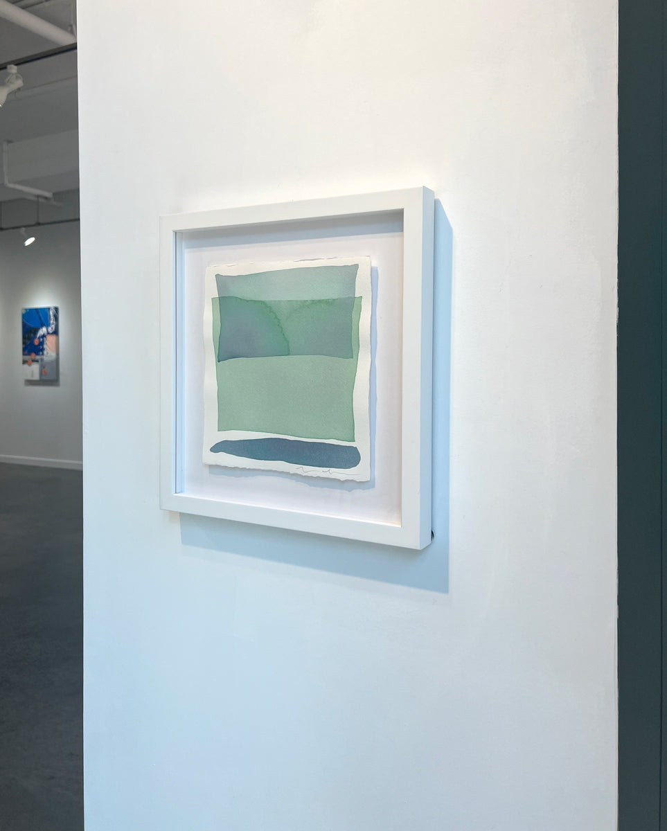 A right side-angle of blue and green shapes overlap in this painting by Nealy Hauschildt in a white frame floating above a white matte hangs on a white wall.