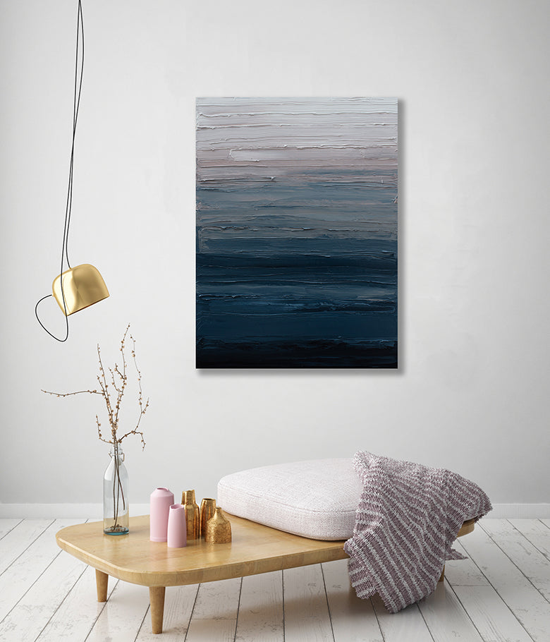 A tonal blue, light pink and white textured abstract painting hangs on a white wall above a decorative ottoman with a cushion and throw blanket.