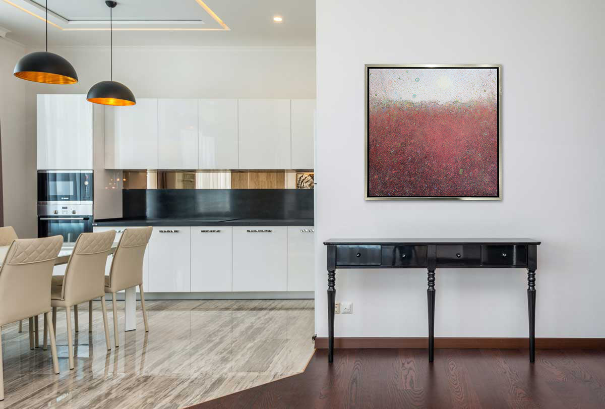 A red abstract landscape print in a silver floater frame hangs on a white wall above a black console table next to an opening of a open kitchen with a a white table and several white chairs.