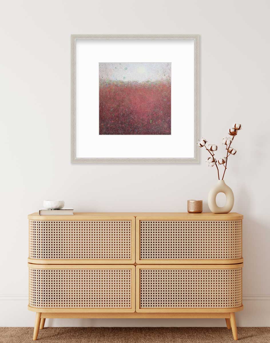 A red abstract landscape print in a silver frame with a mat hangs on a white wall above a wood console cabinet with a book, a white bowl, a copper candleholder and a modern white vase with cotton stems.
