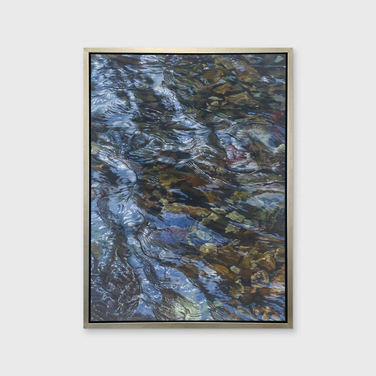 A brown, dark green, white and light blue abstract river rocks in water print in a silver floater frame hangs on a white wall.