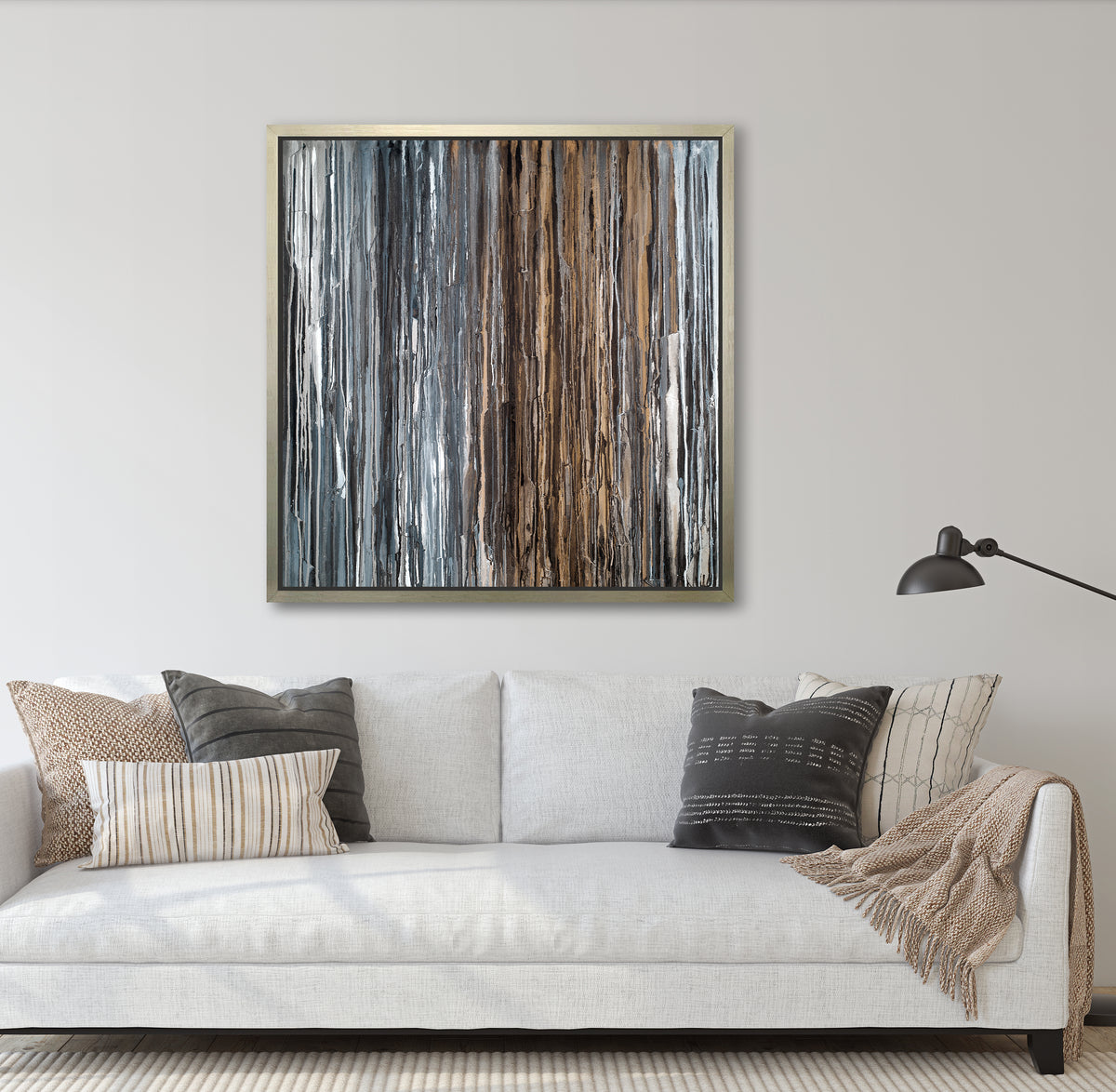 A brown, copper, white, grey and black abstract print in a silver floater frame hangs on a white wall above a white linen sofa with neutral printed pillows and a beige throw blanket.