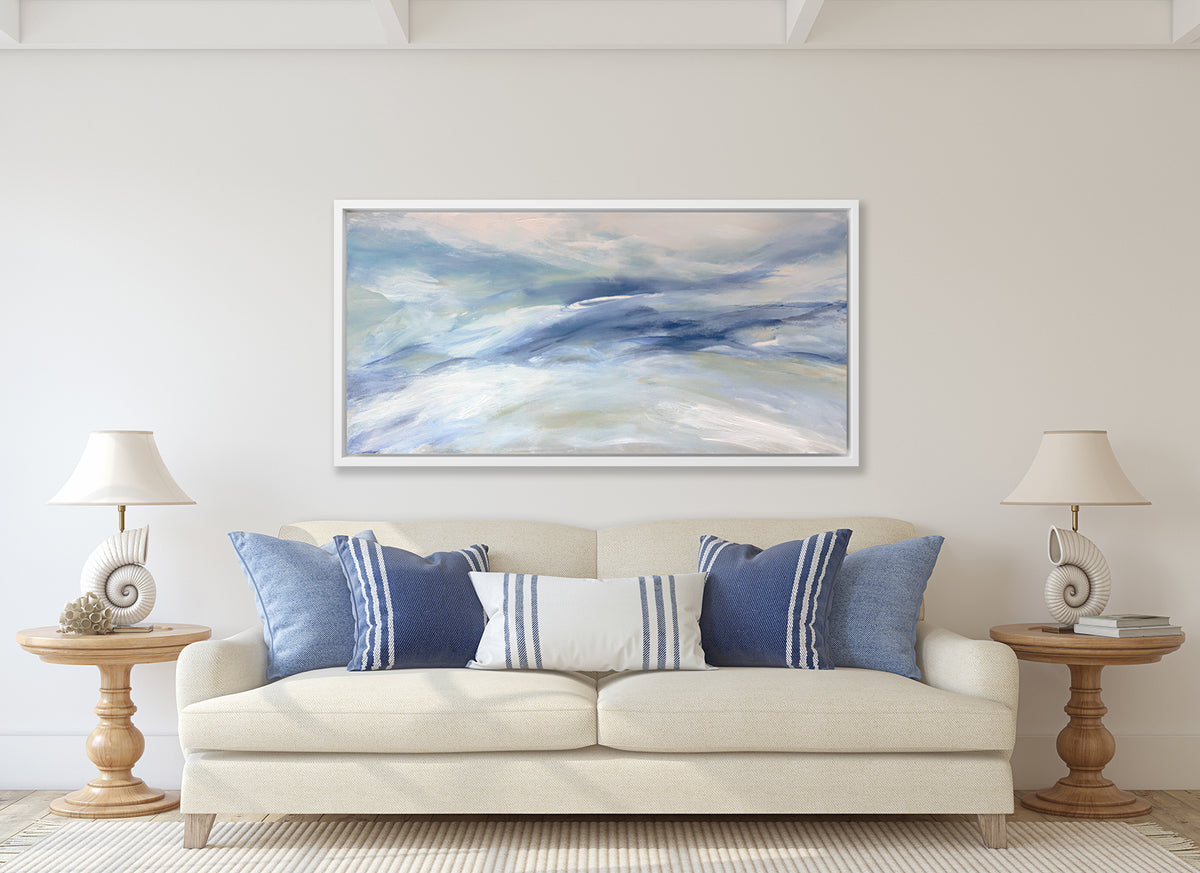 A blue, white and light green abstract print in a white floater frame hangs on a white wall above a cream colored sofa with blue and white pillows, two wood pedestal side tables with matching white seashell table lamps.