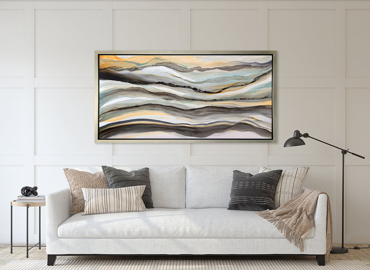 A black, white, orange and teal abstract print in a silver floater frame hangs on a white wall above a white sofa with neutral pillows, a throw blanket and a side table with a stack of books.
