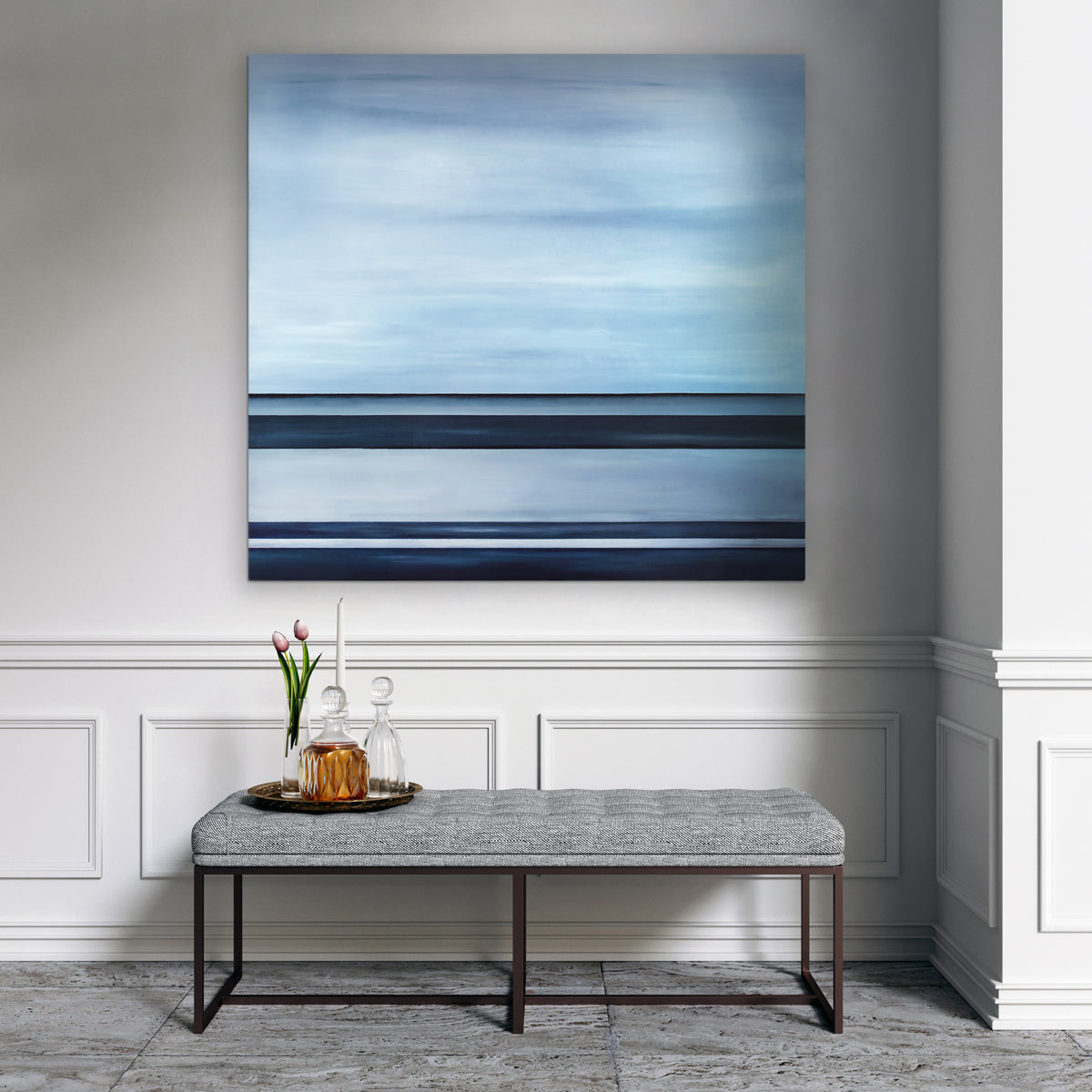A tonal blue linear abstract painting by Tony Iadicicco hangs on a white wall above a grey tufted bench with a tray with two glass decanters, a white tapered candlestick and a glass vase with two pink tulips.