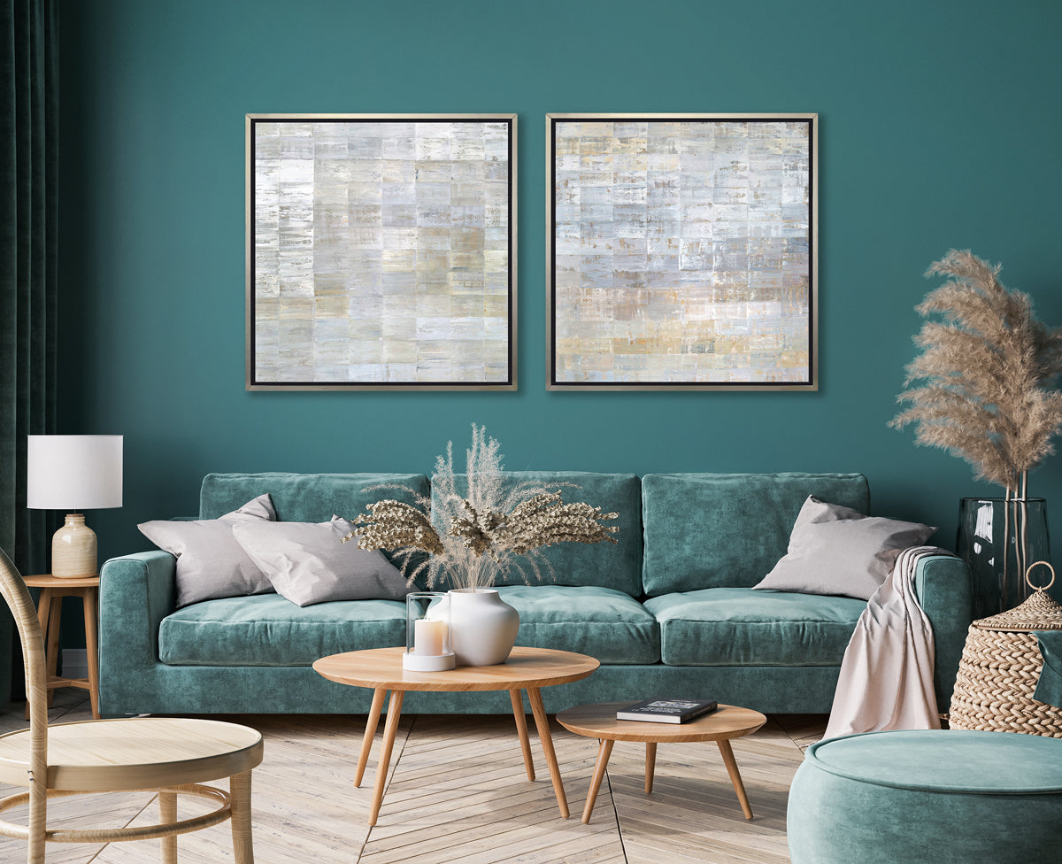 Two grey, silver and light gold abstract geometric prints in silver floater frames hang side by side on a teal wall above a teal sofa with grey pillows, a two tiered round coffee table with a book, a candle and a white vase with floral stems.