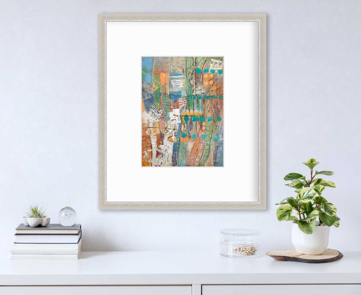 An abstract multicolored print in a silver frame with a mat hangs on a white wall above a white surface with a stack of books, a clear container with gold decorations and a potted plant.