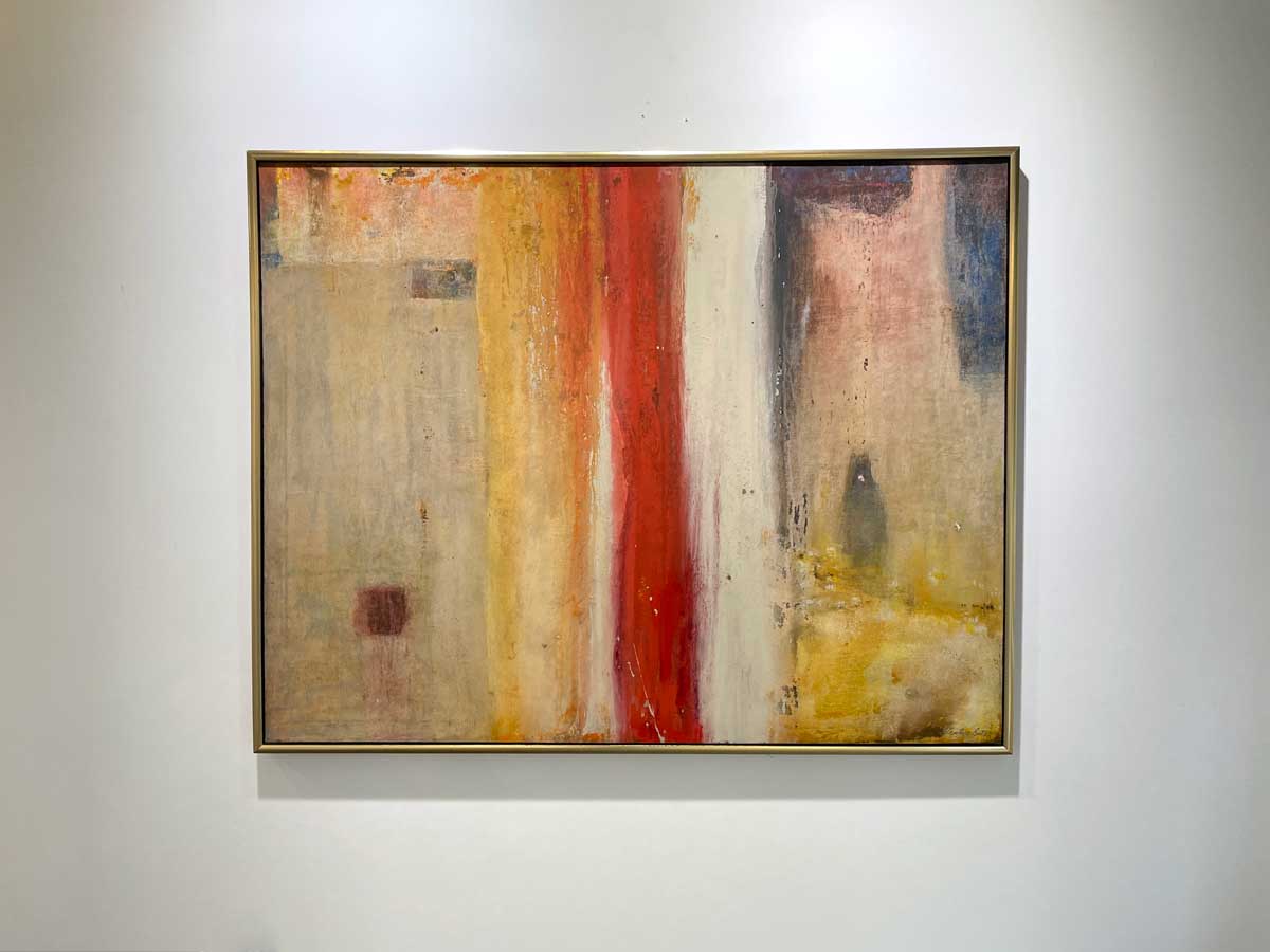 A multicolored abstract painting in a warm gold frame hangs on a white wall.