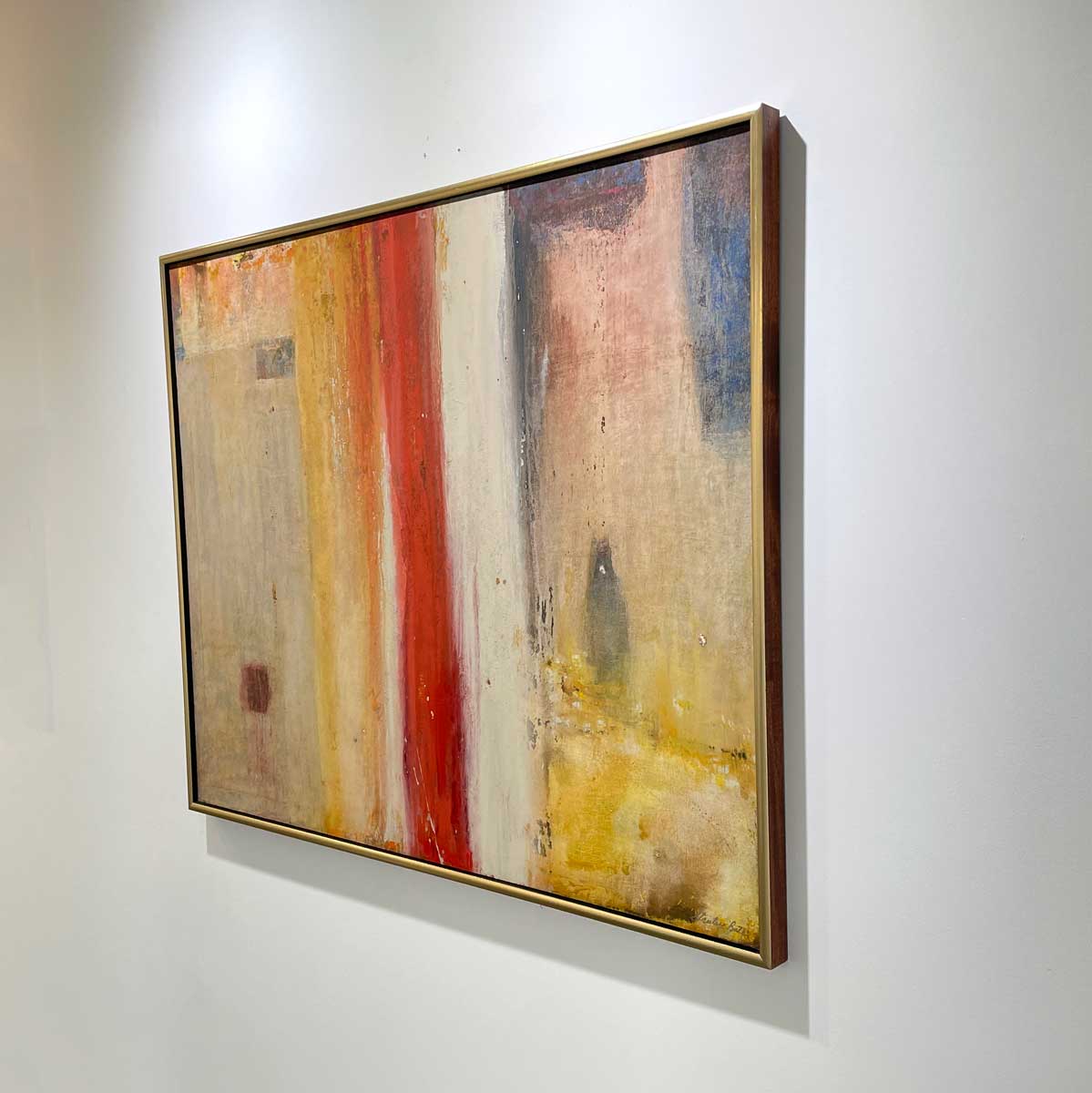 A right side-angle view of a multicolored abstract painting in a warm gold frame hangs on a white wall.