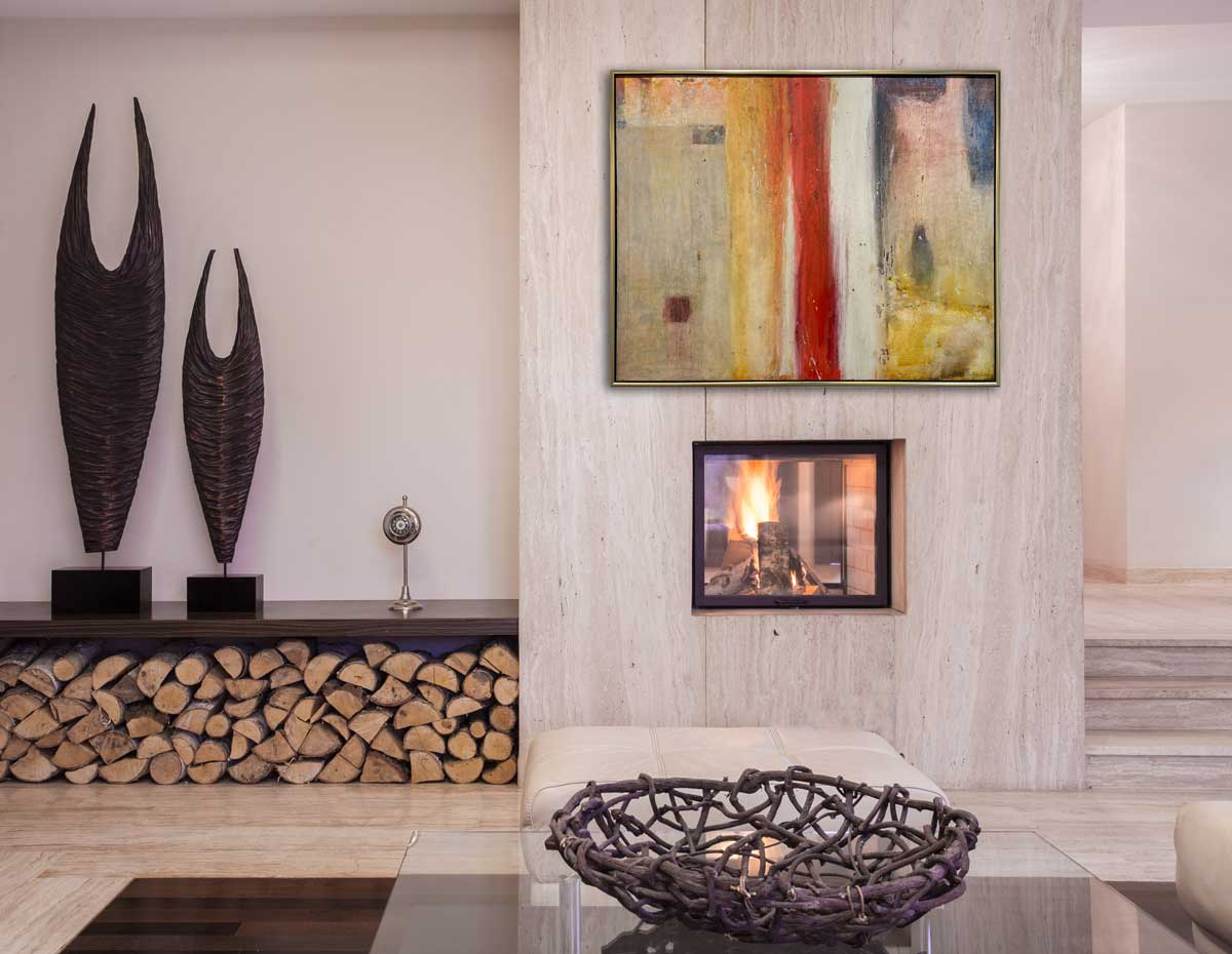 A multicolored abstract painting in a warm gold frame hangs on a white wood wall above a small fireplace insert, a glass coffee table and next to a shelf with two large decorations and wood stacked underneath.