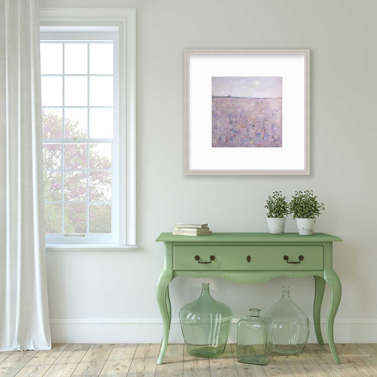 A pink abstract landscape print in a silver frame with a mat hangs on a white wall above a green console table with a stack of books, two potted plants and three green glass bottles.
