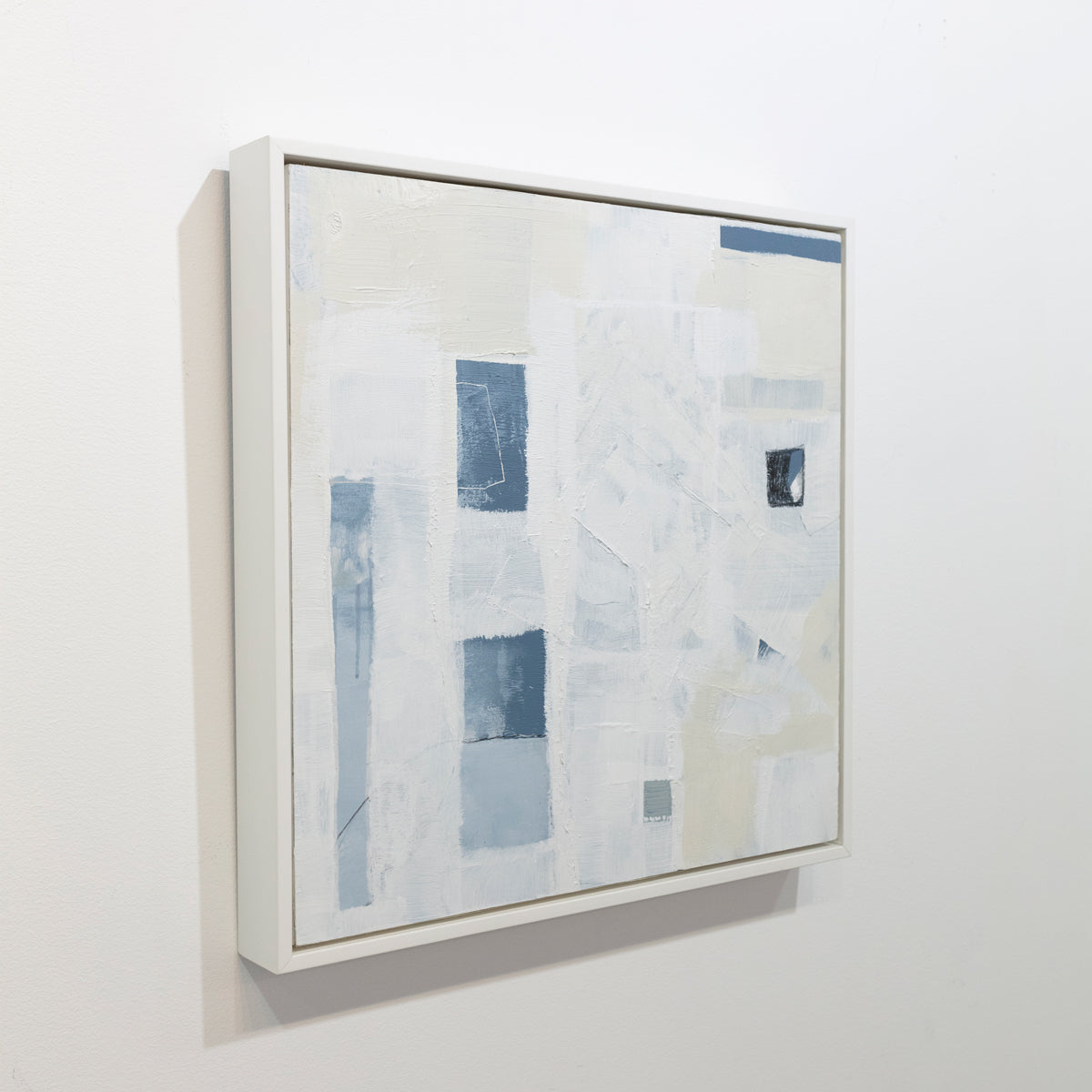 A white and beige abstract painting with blue and black rectangular forms is seen at an angle on a gallery wall. Wired and ready to hang.